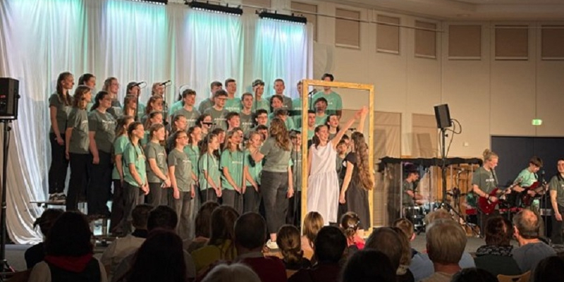 2 junge Frauen stehen sich auf der Bühne spiegelbildlich gegenüber, umrahmt von einem Holzrahmen. Im Hintergrund singt der Chor.