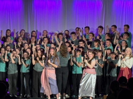 2 junge Frauen tanzen und singen auf der Bühne, im Hintergrund singt der Chor