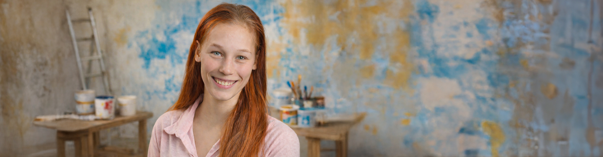 Lächelnde junge Frau mit roten Haaren sitzt vor einer künstlerisch gestalteten Wand mit Farben und Pinseln im Hintergrund.
