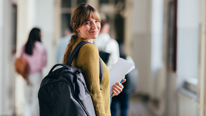Junge Frau mit Rucksack schaut lächelnd über die Schulter in einem Schul- oder Hochschulflur und hält Unterlagen in der Hand.