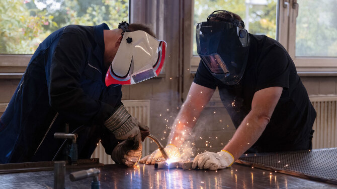 Zwei Personen mit Schutzmasken führen Schweißarbeiten an einem Metallstück in einer Werkstatt durch, Funken sprühen.