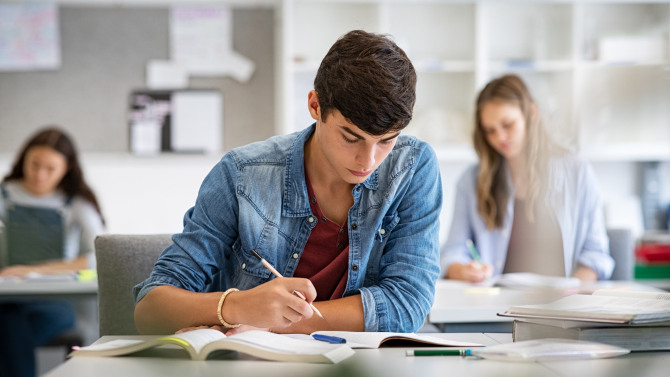 Schüler sitzt konzentriert an einem Tisch und schreibt in ein Buch, während andere im Hintergrund ebenfalls lernen.