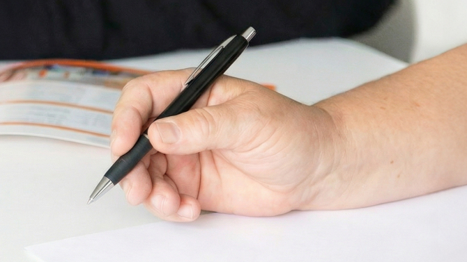Eine Hand hält einen Stift und im Hintergrund liegt eine Broschüre.