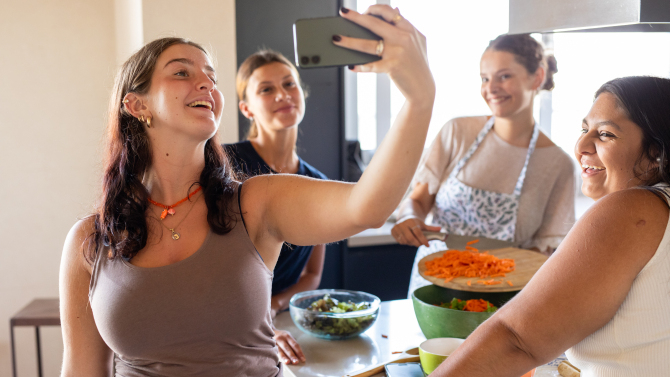 Vier junge Frauen stehen gemeinsam in einer Küche, bereiten Essen zu und machen lachend ein Selfie.