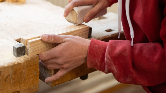 Eine Person schmirgelt ein Holzstück glatt, das in einer Werkbank eingespannt ist.