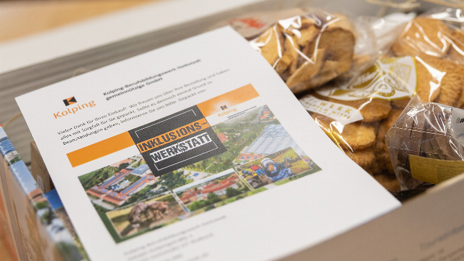 Karton mit Informationsmaterial der Kolping Inklusionswerkstatt neben verpackten Backwaren und Produkten.