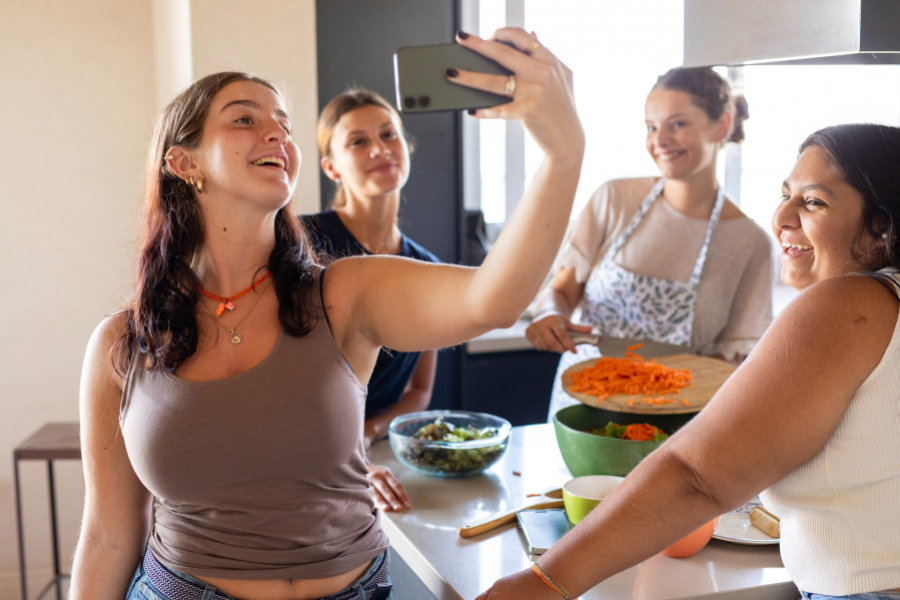 Vier junge Frauen stehen gemeinsam in einer Küche, bereiten Essen zu und machen lachend ein Selfie.