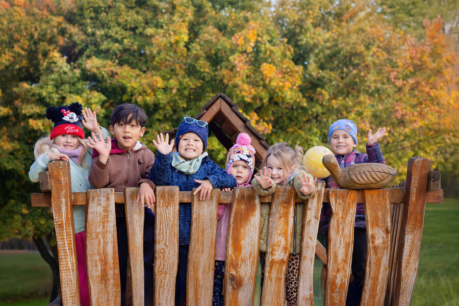 Eine Gruppe von sieben fröhlichen Kindern winkt von einem Holzspielgerät vor herbstlich bunten Bäumen.