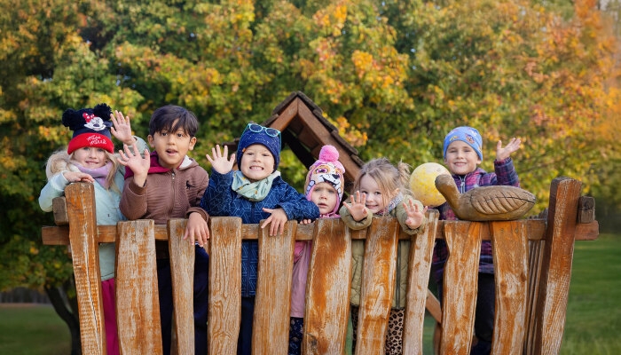 Eine Gruppe von sieben fröhlichen Kindern winkt von einem Holzspielgerät vor herbstlich bunten Bäumen.