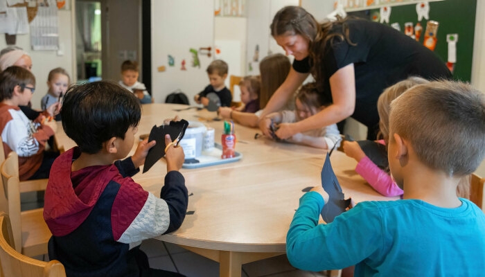 Kinder sitzen an einem Tisch und schneiden schwarze Papierformen aus, eine Erzieherin hilft beim Basteln.