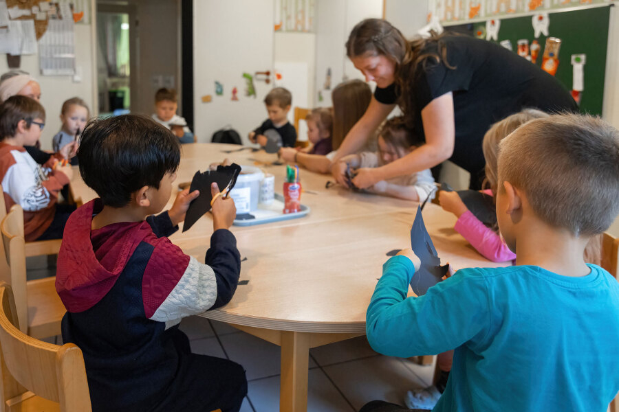 Kinder sitzen an einem Tisch und schneiden schwarze Papierformen aus, eine Erzieherin hilft beim Basteln.