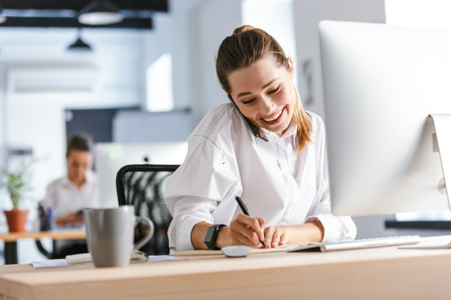Lächelnde Frau sitzt im Büro, telefoniert und macht sich Notizen an ihrem Schreibtisch.