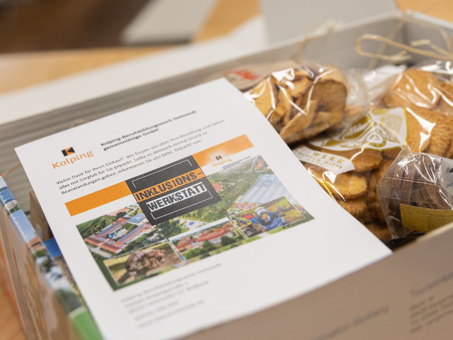 Karton mit Informationsmaterial der Kolping Inklusionswerkstatt neben verpackten Backwaren und Produkten.