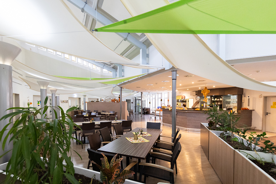 Cafeteria mit Tischen und Stühlen, Theke für Essensausgabe. Zur Deko sind Stoffbahnen an der Decke aufgespannt.