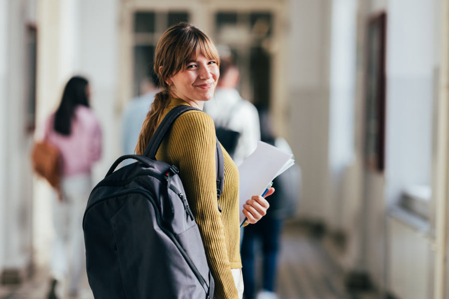 Junge Frau mit Rucksack schaut lächelnd über die Schulter in einem Schul- oder Hochschulflur und hält Unterlagen in der Hand.