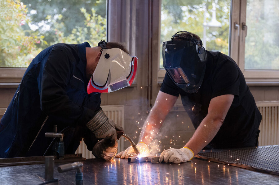 Zwei Personen mit Schutzmasken führen Schweißarbeiten an einem Metallstück in einer Werkstatt durch, Funken sprühen.