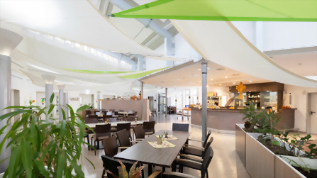 Helle Cafeteria mit Sitzbereichen, hellen Stoffsegeln an der Decke Pflanzen und Ausgabebereich im Kolping Berufsbildungswerk.