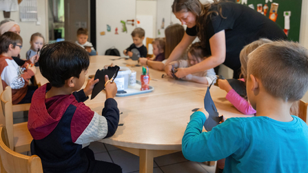 Kinder basteln gemeinsam an einem Tisch mit einer p&auml;dagogischen Fachkraft in einem Gruppenraum.