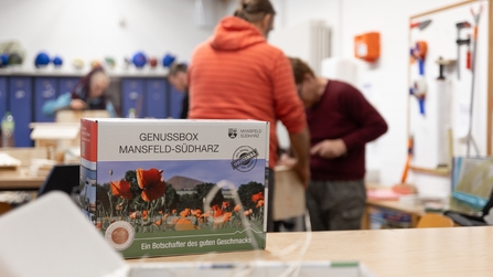 Fertige Genussbox steht im Vordergrund, im Hintergrund arbeiten Menschen der Inklusionswerkstatt